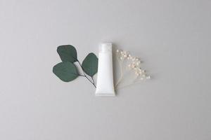 vit rör av kosmetisk grädde med blommor och grön löv på grå bakgrund. platt lägga, kopia Plats foto