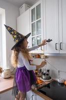 barn dekorerar de kök i Hem för halloween. flicka i en häxa kostym spelar med de dekor för de Semester - fladdermöss, domkraft lykta, pumpor. höst bekvämlighet i hus, scandi-stil kök, loft foto