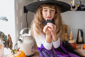 barn dekorerar de kök i Hem för halloween. flicka i en häxa kostym spelar med de dekor för de Semester - fladdermöss, domkraft lykta, pumpor. höst bekvämlighet i hus, scandi-stil kök, loft foto