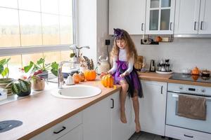 barn dekorerar de kök i Hem för halloween. flicka i en häxa kostym spelar med de dekor för de Semester - fladdermöss, domkraft lykta, pumpor. höst bekvämlighet i hus, scandi-stil kök, loft foto