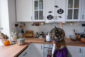 barn dekorerar de kök i Hem för halloween. flicka i en häxa kostym spelar med de dekor för de Semester - fladdermöss, domkraft lykta, pumpor. höst bekvämlighet i hus, scandi-stil kök, loft foto