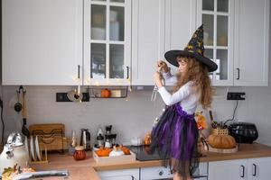 barn dekorerar de kök i Hem för halloween. flicka i en häxa kostym spelar med de dekor för de Semester - fladdermöss, domkraft lykta, pumpor. höst bekvämlighet i hus, scandi-stil kök, loft foto