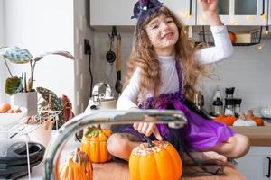 barn dekorerar de kök i Hem för halloween. flicka i en häxa kostym spelar med de dekor för de Semester - fladdermöss, domkraft lykta, pumpor. höst bekvämlighet i hus, scandi-stil kök, loft foto