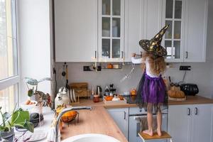 barn dekorerar de kök i Hem för halloween. flicka i en häxa kostym spelar med de dekor för de Semester - fladdermöss, domkraft lykta, pumpor. höst bekvämlighet i hus, scandi-stil kök, loft foto