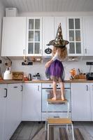 barn dekorerar de kök i Hem för halloween. flicka i en häxa kostym spelar med de dekor för de Semester - fladdermöss, domkraft lykta, pumpor. höst bekvämlighet i hus, scandi-stil kök, loft foto