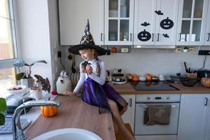 barn dekorerar de kök i Hem för halloween. flicka i en häxa kostym spelar med de dekor för de Semester - fladdermöss, domkraft lykta, pumpor. höst bekvämlighet i hus, scandi-stil kök, loft foto