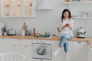 kvinna hemmafru nöjd med hus rengöring tjänster sitter i modern rena kök med smartphone foto