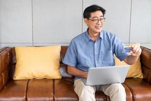 inomhus- skott inuti av de hus av senior asiatisk män sitta ner på soffa och använder sig av dator bärbar dator för uppkopplad kreditera kort betalning med leende på hans ansikte. Lycklig senior med teknologi. foto