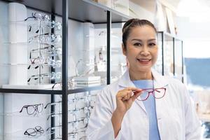 asiatisk kvinnor läkare optiker leende medan använder sig av läsplatta och rekommendera kund till välja glasögon skön ram rättegång inuti av optisk affär, Lagra. öga syn för patient, sjukvård begrepp. foto