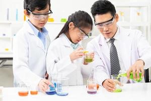 ung asiatisk barn bär vit forskare klänning och do kemisk testa rör medan studie, inlärning i vetenskap klassrum med lärare. ung barn utbildning begrepp förbi experimentera, roligt och njut av klassrum foto