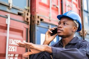 selektiv fokus på ansikte av svart afrikansk man arbetstagare bär säkerhet Utrustning, talande på smartphone medan kolla upp och inspektion de tillstånd av behållare. importera och exportera, logistisk frakt arbetstagare. foto