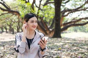 selektiv fokus på ansikte av ung skön asiatisk kvinnor använder sig av smart Kolla på till Spår aktivitet och lyssna musik från smartphone medan värma upp innan övning yoga med naturlig och träd på bakgrund. foto