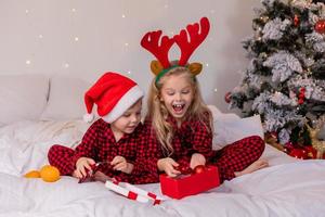 två Lycklig barn i pyjamas för jul är sortering presenterar och spelar med jul leksaker. hög kvalitet Foto