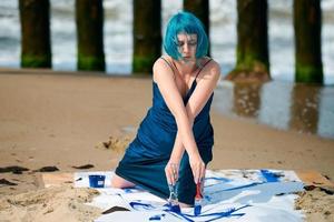 konstnärlig blåhårig kvinna performancekonstnär insmord med gouachefärger på stor duk på stranden foto