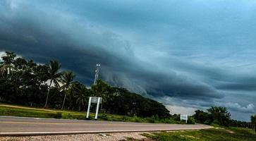 svart storm moln form över en svart väg omgiven förbi grön skogar. monsun säsong väder prognos. foto