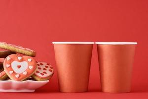 dekorerad hjärta form småkakor och två papper kaffe koppar på de röd bakgrund. valentines dag mat begrepp foto