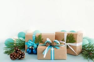 tre jul gåva lådor insvept av hantverk papper, blå och vit band, dekorerad av gran grenar, tall koner och jul bollar på de blå och vit bakgrund med jul lampor. foto