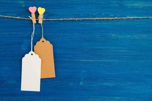 två brun och vit tom papper pris taggar eller etiketter uppsättning och trä- stift dekorerad på hjärtan hängande på en rep på de blå trä- bakgrund. foto