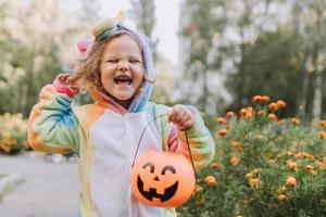 söt liten flicka i en regnbåge enhörning kostym för halloween går till samla sötsaker i en pumpa korg i en bostads- område. barn promenader i de utomhus. lura eller behandla. livsstil. kigurumi foto