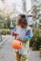 söt liten flicka i en regnbåge enhörning kostym för halloween går till samla sötsaker i en pumpa korg i en bostads- område. barn promenader i de utomhus. lura eller behandla. livsstil. kigurumi foto