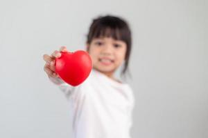 de liten flicka i en vit skjorta innehav en röd hjärta på en vit bakgrund. hälsning kort för hjärtans dag, mors dag, fars dag. foto