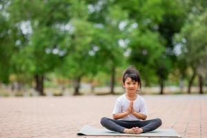 liten söt asiatisk flicka praktiserande yoga utgör på en matta i parkera, friska och övning begrepp foto