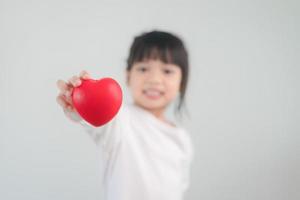 de liten flicka i en vit skjorta innehav en röd hjärta på en vit bakgrund. hälsning kort för hjärtans dag, mors dag, fars dag. foto