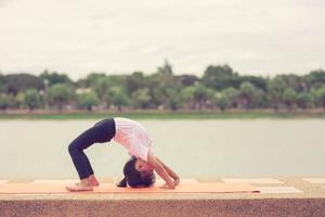 liten söt asiatisk flicka praktiserande yoga utgör på en matta i parkera, friska och övning begrepp foto