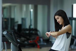 en liten flicka utseende på smart Kolla på efter Träning i Gym. friska begrepp. kvinna efter träna session checkar resultat på titta.barns sport begrepp foto