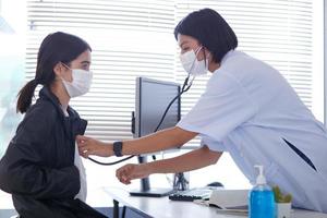 ett asiatisk kvinna läkare bär en kirurgisk mask är kontroll de barn patientens hjärta Betygsätta med en stetoskop. foto