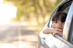 söt asiatisk liten barn flicka har roligt till resa förbi bil och ser ut från ar fönster i cthe landsbygden foto