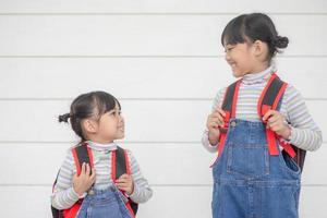 tillbaka till skola. två söt asiatisk barn flickor med skola påsar innehav en bok tillsammans på vit bakgrund foto