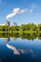 rör av träbearbetning företag växt sågverk med skön reflexion i blå vatten av flod. luft förorening begrepp. industriell landskap miljö- förorening avfall av termisk kraft växt foto