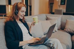 upphetsad rödhårig leende kvinna med trådlöst headset och laptop sitter på soffan i caféet foto