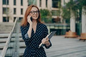 positiv rödhårig europeisk kvinna har telefonsamtal medan du går utomhus foto