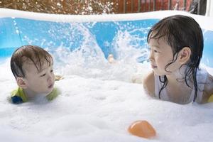 söt asiatisk syskon har roligt i de simning slå samman. syster och bror spelar med bubblor och simning i de sommar semester slå samman. Lycklig familj Semester begrepp foto