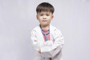 ung söt asiatisk pojke bär en medicinsk enhetlig innehav en stetoskop spelar Lycklig läkare på vit bakgrund. förskola barn låtsas till vara en barnläkare. foto