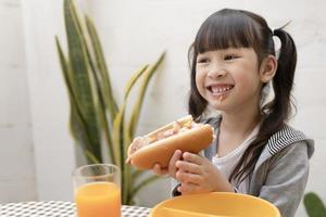 ung asiatisk flicka äter frukost och orange juice på de tabell på Hem fortfarande Lycklig. friska äter, mat och mellanmål, skinka och ost smörgås, barn Lycklig äter foto