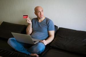 emotionell skallig man på soffa med bärbar dator och kreditera kort i hand foto