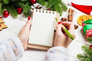 topp se av kvinna hand skrivning i en anteckningsbok på trä- jul bakgrund. gran träd och festlig dekorationer. önskar lista. ny år begrepp foto