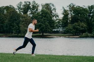 stilig atlet skäggig man springer utomhus på morgonen poser nära floden njuter av naturen och frisk luft, visar uthållighet och motivation, tränar på morgonen varje dag, håller sig frisk och stark foto