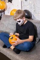 en pojke i skelett smink målarfärger en pumpa för halloween. foto