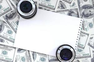 två fotografisk linser och vit anteckningsbok lögn på de bakgrund av en massa av dollar räkningar. Plats för text foto