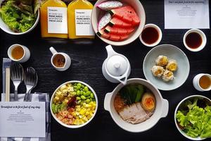 ovanifrån av frukost med färsk sallad, dim sum, nudlar med kokt fläsk, vattenmelon, drakfrukt, flaska apelsinjuice och söt sås. platt låg av hälsosam mat med grönsaker och frukt på bordet. foto