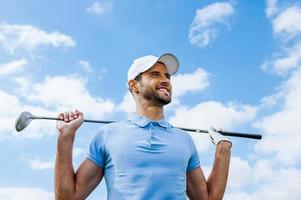 ser mot Framgång. låg vinkel se av ung Lycklig golfspelare innehav förare och leende med blå himmel som bakgrund foto