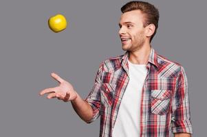 jonglering hans friska livsstil. en leende man kasta ett äpple in i de luft medan stående mot grå bakgrund foto