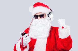 njuter jul musik. santa claus i solglasögon och hörlurar lyssnande till mp3 spelare och gestikulerar medan stående mot grå bakgrund foto