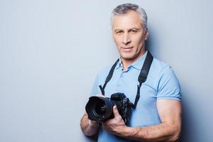 expert- i fotografi. porträtt av senior mogna man i t-shirt innehav kamera medan stående mot grå bakgrund foto