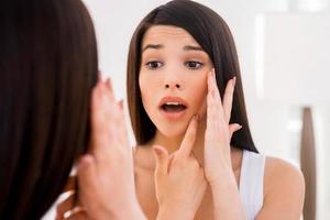orolig handla om acne. frustrerad ung kvinna granskning henne ansikte medan ser på de spegel foto