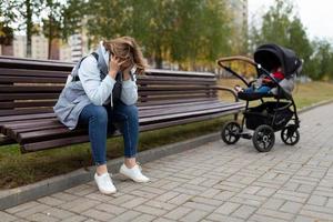 en ung mor sitter på en bänk upprörd Nästa till en bebis sittvagn, begrepp av postpartum depression foto